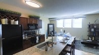 This single-floor space showcases an open-concept layout that seamlessly connects the kitchen and living area, creating a cohesive and functional environment. The kitchen is anchored by rich, dark wood cabinetry that extends to the upper walls, complemented by a classic tile backsplash and black appliances, including a range and over-the-range microwave. A spacious granite-topped island serves as the centerpiece, doubling as a breakfast bar with seating, and features a stainless steel sink with a modern arc faucet. The light-toned granite countertops provide a striking contrast against the darker cabinetry. Beyond the kitchen, the open living and dining area benefits from large windows that invite generous natural light, enhancing the bright, airy atmosphere of the space. A dining table positioned near the windows offers a well-lit area for meals, while the adjoining living space provides room for comfortable seating. A full-length mirror near the living area further amplifies the sense of openness. Overhead, a prominent circular flush-mount light fixture illuminates the kitchen, while industrial-style pendant lighting accents the living area, adding a contemporary touch throughout.