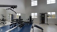 This fitness center is designed to provide a functional and motivating environment for exercise. The space is defined by soaring ceilings that create an open, airy atmosphere, while multiple windows positioned at both standard and elevated heights flood the room with natural light and offer views of the surrounding outdoor scenery. The walls are finished in a clean, neutral gray, contributing to a focused and professional ambiance. Large mirrors line one wall, enhancing the sense of space and allowing users to monitor their form during workouts. The flooring transitions between a durable blue rubber mat in the primary exercise zone and a light-colored hard surface, providing both stability and practicality. Equipment includes a full dumbbell rack with a wide range of weights, a dedicated weight bench, a plate-loaded strength machine, and a multi-functional cable machine, offering a thorough selection of options for strength training and resistance work. The layout is thoughtfully organized to maximize usable floor space while maintaining clear pathways between stations. Overall, the fitness center delivers a clean, bright, and well-appointed setting suited for a comprehensive strength-focused workout routine.