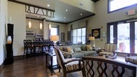 This single-floor space showcases an open-concept design that seamlessly connects the lounge and dining areas, creating a welcoming and functional environment. Soaring ceilings with dark crown molding and recessed lighting add a sense of grandeur, while pendant lights contribute warm, layered illumination throughout the space.

The living area is anchored by a neutral-toned sofa accented with decorative pillows, complemented by stylish arm chairs featuring intricate lattice-patterned frames. A sleek coffee table sits atop a patterned area rug, bringing cohesion to the seating arrangement. Wall art and tall decorative branches add artistic focal points throughout.

The adjacent dining area features a bar-height table surrounded by matching chairs with ornate detailing, positioned near a kitchen counter with appliances visible in the background. Dark hardwood flooring runs throughout, tying the two zones together elegantly.

Natural light filters in through multiple windows dressed with layered curtains, brightening the neutral beige walls. The overall ambiance is sophisticated yet inviting, blending contemporary furnishings with classic architectural details to create a polished, well-appointed space.