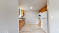 This single-floor unit offers a practical and well-organized layout designed for comfortable everyday living. The kitchen is equipped with warm honey-toned wood cabinetry, granite-look countertops, a double sink, and white appliances including a refrigerator, range, microwave, and dishwasher. Light wood-look vinyl flooring runs throughout the kitchen, providing a clean and easy-to-maintain surface.

Adjacent to the kitchen, the living area transitions to neutral wall-to-wall carpeting, creating a softer, more comfortable atmosphere. A ceiling fan and windows fitted with horizontal blinds allow for natural light and airflow. Baseboard heating units are present throughout the unit, ensuring consistent warmth.

The bedrooms feature the same plush carpeting, with windows overlooking greenery outside. Each bedroom includes a closet with a hanging rod and shelf for organized storage. One bedroom benefits from direct access to the bathroom, adding a degree of convenience.

The bathroom is finished with white tile wainscoting, a single-sink vanity with wood cabinetry, a full soaking tub, and tile flooring. A window with blinds provides ventilation and natural light. The overall aesthetic of the unit is clean, neutral, and move-in ready.