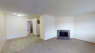This single-floor space offers a functional and straightforward layout connecting the main living area to the kitchen and additional rooms beyond. The living area is finished with wall-to-wall carpeting and features a notable stone-surround fireplace as a focal point, adding a classic, cozy element to the room. The textured popcorn ceiling runs throughout, contributing to the space's traditional character.

Transitioning from the carpeted living area, the flooring shifts to tile as you move toward the kitchen zone, clearly delineating the two areas. The kitchen is visible from the living space and includes warm-toned wooden cabinetry and a white refrigerator, providing a practical and straightforward cooking environment. A hallway extends beyond, leading to additional rooms and suggesting a well-connected floor plan. The walls throughout are finished in neutral, light tones, creating a clean and versatile backdrop. Overall, the space presents a practical, classic interior with a comfortable, lived-in character suited for everyday use.
