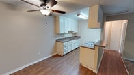 This single-floor residence offers a practical and comfortable layout designed for everyday living. The kitchen is a functional galley-style space featuring white upper and lower cabinetry, dark laminate countertops, a double basin sink, a gas range with oven, and durable wood-look flooring. A peninsula counter with a raised breakfast bar separates the kitchen from the adjoining living and dining area, creating an open, connected feel. A ceiling fan with a light fixture overhead provides both airflow and ambient lighting throughout the combined space.

The living area transitions seamlessly between wood-look flooring and plush carpeting, offering a cozy and versatile environment. A sliding glass door opens to a private balcony, allowing natural light to filter in and providing pleasant outdoor views of surrounding greenery.

The bedroom is generously proportioned, featuring soft carpet flooring, neutral-toned walls, a textured ceiling, and a window dressed with vertical blinds that admit natural light while maintaining privacy.

The bathroom is clean and straightforward, with a combined tub and shower unit, a toilet, and a vanity with a light countertop. Wood-look flooring ties the space together with the rest of the unit. Throughout, the home presents a neutral color palette, textured ceilings, and consistent finishes that create a cohesive and move-in-ready atmosphere.