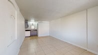 This single-floor space offers a straightforward, functional layout comprising an open living area, a built-in kitchenette, and a full bathroom. The main living area is finished with large-format light beige ceramic tile flooring, crisp white walls, and a textured popcorn ceiling, creating a clean and neutral interior. A wall-mounted PTAC heating and cooling unit provides climate control, while a front-facing window with horizontal blinds allows natural light to filter in and offers a street-level view. The entry door is positioned adjacent to this window, providing direct access to the space.

The kitchenette is integrated into one corner of the main room and features gray cabinetry, a dark countertop, a sink, and an overhead microwave, offering essential cooking and preparation amenities in a compact footprint. A mirrored cabinet above the sink adds a practical touch.

The bathroom is a separate enclosed room, finished with large-format gray tile on both the floors and walls. It includes a walk-in shower with a chrome showerhead, a toilet, and a vanity with a dark granite-style countertop. The consistent tile work throughout the bathroom gives it a clean, modern appearance. Overall, the space is efficiently designed, making practical use of every square foot available.