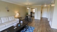 This single-floor residence offers a thoughtfully designed open-concept layout that combines the living, dining, and kitchen areas into one cohesive and functional space. Rich hardwood-style flooring runs throughout, lending warmth and continuity to the interior. The living area is comfortably sized, featuring a neutral color palette on the walls that is complemented by elegant crown molding, giving the space a polished, finished look. A dark coffee table and plush seating anchor the lounge area, while a vibrant blue area rug adds a pop of color and visual interest. Adjacent to the living space, the dining area accommodates a round wooden table with industrial-style bar stools beneath a classic pendant light fixture, creating an inviting spot for meals and gatherings. The kitchen is accessible through a wide doorway, revealing white cabinetry, dark countertops, and a window that draws in natural light. A hallway extending from the main living area leads to additional rooms, maintaining a clear and efficient flow throughout the home. The overall ambiance is warm, clean, and welcoming, with a balance of modern and classic design elements throughout.