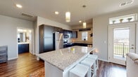 This thoughtfully designed single-floor residence offers a seamless blend of modern finishes and comfortable living spaces. Rich hardwood-style flooring flows throughout much of the home, creating a cohesive and inviting atmosphere from entry to living area.

The kitchen serves as a central highlight, showcasing dark cabinetry contrasted by light granite countertops, stainless steel appliances, and a large island with bar seating. Pendant lighting overhead adds a polished touch, while the adjoining dining nook provides an intimate space for everyday meals.

The living area is bathed in natural light from large windows and offers direct access to an outdoor balcony, framing pleasant neighborhood views. The bedrooms are carpeted for comfort, each featuring windows that bring in ample daylight and outdoor scenery. The primary bedroom is generously proportioned and connects to additional private spaces.

A full bathroom includes a tub-shower combination and continues the home's consistent use of wood-look flooring. The dedicated laundry room is equipped with a stacked washer and dryer unit alongside built-in storage drawers, maximizing functionality. The entryway corridor is practical and welcoming, with additional storage cabinetry near the front door. Throughout the home, neutral tones and clean architectural lines create a calm, move-in-ready environment.