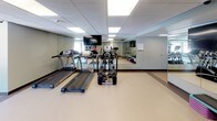 This fitness center is a thoughtfully designed, single-floor facility offering a comprehensive range of exercise equipment suited for various workout styles. The space is organized into distinct functional zones while maintaining an open, cohesive layout throughout.

The cardio area features treadmills and elliptical trainers positioned alongside full-wall mirrors that amplify the sense of space. A wall-mounted flat-screen television provides entertainment during workouts. Recessed panel lighting in the drop ceiling ensures consistent, even illumination across the entire floor.

An adjoining zone houses upright and recumbent stationary bikes alongside aerobic step platforms, catering to low-impact training needs. A water station is conveniently accessible nearby. Large windows in this section introduce abundant natural light and offer views of a wooded outdoor landscape.

The strength training area is well-appointed with multi-station cable machines, a rowing machine, and dedicated floor space for functional movements. A freestanding rack organizes stability balls, medicine balls, and kettlebells of varying weights, supporting dynamic and core-focused training.

The flooring throughout is durable, easy-to-clean vinyl in neutral tones with contrasting border accents. Artwork displayed on the walls adds a welcoming, polished atmosphere to the overall space.