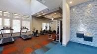 The fitness center at Apalachee Point is a thoughtfully designed workout facility that combines functionality with contemporary style. The space features soaring ceilings that create an open, airy atmosphere, enhanced by recessed lighting and a ceiling fan that ensure a comfortable environment during workouts. A striking mosaic tile accent wall in cool blue tones adds a bold visual element, complementing the vibrant, multicolored carpet tiles that define the exercise floor — a dynamic mix of orange, teal, and gray patterns that energize the space. Natural light filters in through multiple windows fitted with blinds, keeping the area bright and welcoming. The equipment layout is well-organized, offering a variety of cardio machines including treadmills, ellipticals, and stationary bikes, alongside a multi-use strength training station. A dedicated mirrored section provides an ideal space for strength work and form checks. The ceiling transitions between open, lofted areas and a coffered section that adds architectural interest. Overall, the fitness center at Apalachee Point delivers a clean, modern, and fully equipped environment suitable for a comprehensive range of fitness routines.
