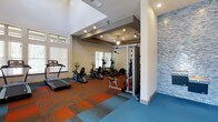 The fitness center at Apalachee Point is a thoughtfully designed workout facility that combines functionality with contemporary style. The space features soaring ceilings that create an open, airy atmosphere, enhanced by recessed lighting and a ceiling fan that ensure a comfortable environment during workouts. A striking mosaic tile accent wall in cool blue tones adds a bold visual element, complementing the vibrant, multicolored carpet tiles that define the exercise floor — a dynamic mix of orange, teal, and gray patterns that energize the space. Natural light filters in through multiple windows fitted with blinds, keeping the area bright and welcoming. The equipment layout is well-organized, offering a variety of cardio machines including treadmills, ellipticals, and stationary bikes, alongside a multi-use strength training station. A dedicated mirrored section provides an ideal space for strength work and form checks. The ceiling transitions between open, lofted areas and a coffered section that adds architectural interest. Overall, the fitness center at Apalachee Point delivers a clean, modern, and fully equipped environment suitable for a comprehensive range of fitness routines.