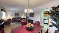 Located at 98 Oak Street in Lindenwold, New Jersey, this single-floor home offers a practical and welcoming layout designed for comfortable everyday living. The open-concept main living area seamlessly connects the dining and living spaces, creating a unified environment that feels both spacious and inviting. A statement chandelier with a classic brushed-nickel finish serves as a focal point above the dining area, casting warm light over the rich, dark-toned dining table and chairs. The living room is anchored by a neutral-toned sofa set arranged around a rectangular coffee table, with a wall-mounted flat-screen television providing a modern touch. Soft carpeting throughout the main living area adds warmth underfoot, while neutral-painted walls contribute to the bright and airy atmosphere. Adjacent to the main living space, the kitchen is accessible through an open passageway, featuring wooden cabinetry, tile flooring, and essential appliances, making it both functional and easy to navigate. Decorative accents, including framed artwork on the walls and a decorative bowl centerpiece on the dining table, add personality and a lived-in charm to the home. Overall, this residence offers a cohesive, single-level floor plan well-suited for straightforward, comfortable living.