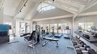 This single-floor fitness center is designed to support a full range of workout routines in a bright, open environment. The space is defined by its striking vaulted ceiling with exposed white beam detailing, creating a sense of impressive height and volume throughout. A large ceiling fan anchors the apex of the ceiling, complementing recessed lighting and track lighting to ensure the space is well-lit at all times.

Natural light floods the interior through an array of large windows lining multiple walls, offering views of the outdoor pool area and surrounding greenery. The blue patterned carpet flooring ties the space together while providing comfort underfoot.

The fitness center is comprehensively equipped with a wide variety of cardio machines, including multiple treadmills, ellipticals, and stationary bikes, all positioned along the windowed walls to maximize outdoor views during workouts. The strength training area features cable machines, a Smith machine, weight benches, barbells, and a full dumbbell rack. Full-length mirrors along one wall enhance the sense of space and allow for form monitoring during exercise.

Additional amenities include a stretching and mat area with foam rollers and fitness accessories. The facility is accessible via glass-paned double doors, adding to the welcoming, airy ambiance of this well-appointed community fitness space.