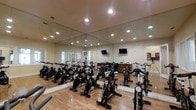 This purpose-built spinning room is designed to deliver a focused and energizing indoor cycling experience. The space is outfitted with a full arrangement of high-quality stationary spin bikes, positioned in rows across a warm hardwood floor that adds both durability and visual warmth to the environment. Floor-to-ceiling mirrors line the walls, creating a sense of expanded space while allowing participants to monitor their form throughout their workout. Recessed lighting is evenly distributed across the ceiling, providing bright, consistent illumination throughout the room. Multiple wall-mounted flat-screen displays are positioned for clear sightlines from the bikes, ideal for instructor-led classes or visual content during sessions. A neutral, light-toned color palette on the walls and ceiling contributes to a clean and focused atmosphere. The room also features a camera or recording stand near the front, suggesting the space is equipped to support virtual or live-streamed classes. Entry is accessible via a door visible in the mirror's reflection, connecting seamlessly to adjacent amenity areas. Overall, the spinning room is a well-appointed, fully functional fitness space thoughtfully designed to accommodate group cycling classes at a comfortable capacity.