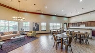 This single-floor community space is designed with an open-concept layout that seamlessly integrates a lounge area, dining space, and a fully equipped kitchen. Soaring ceilings with dark crown molding lend an air of grandeur throughout, while large windows line the walls, flooding the interior with abundant natural light and offering views of the surrounding outdoor areas.

The lounge area is appointed with a tufted settee, accent chairs, and a bold equestrian-themed wall art piece, all anchored by a patterned area rug beneath a striking circular chandelier with exposed Edison bulbs. Decorative accent tables and sculptural elements complete the refined yet welcoming atmosphere.

The dining area features multiple dark-metal table-and-chair sets arranged across warm wood-look flooring, providing ample seating for gatherings and events. A built-in granite-topped bar counter with pendant lighting serves as a central focal point, ideal for entertaining.

The kitchen is outfitted with rich dark cabinetry, granite countertops, a classic white subway tile backsplash, stainless steel appliances including a refrigerator and dishwasher, and a double-basin sink. Bar-height seating along the kitchen island adds additional functionality. The overall aesthetic is contemporary and inviting, blending practical amenities with stylish design details throughout.