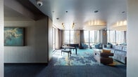 This elegant, single-floor high-rise amenity space combines refined design with a variety of functional zones, all unified by a striking teal, navy, and gold abstract-patterned carpet that anchors the interior. Floor-to-ceiling windows wrap around the space, flooding it with abundant natural light and offering sweeping panoramic views of the surrounding cityscape and skyline beyond.

The main lounge area is a standout, featuring a generously sized tufted gray sectional sofa complemented by teal accent chairs and a low-profile coffee table. Overhead, large sculptural drum pendant lights hang from a high, polished ceiling, adding warmth and a sense of grandeur. Gold cylindrical pendant fixtures provide additional accent lighting throughout.

A full-size black grand piano positioned near the windows serves as a focal point, underscoring the space's upscale character. Adjacent to the lounge, a dedicated meeting or co-working area accommodates a long conference table surrounded by dark chairs, ideal for collaborative work sessions with a dramatic city backdrop.

The entry vestibule features dark-toned walls and tiled flooring, providing a transitional buffer from the elevator lobby. A built-in media console with a flat-screen display and decorative shelving adds functionality to the lounge. Throughout, the design palette of charcoal, gold, and teal creates a cohesive, contemporary atmosphere suited for both social gatherings and productive work.