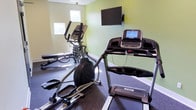 This fitness center offers a focused and functional workout environment designed to support a well-rounded exercise routine. The space is finished with a vibrant two-tone color palette — a bold lime green accent wall paired with soft cream-colored walls — creating an energizing yet clean atmosphere conducive to physical activity. Durable rubber flooring covers the entire area, providing traction and cushioning ideal for a variety of exercises.

The cardio section is equipped with a Spirit treadmill and an elliptical trainer, both positioned to face a wall-mounted flat-screen television, allowing users to stay entertained during their workouts. Recessed lighting throughout the ceiling ensures the space is well-lit, while windows bring in natural light and provide views to the outdoors, enhancing the overall sense of openness.

The strength training area includes a full dumbbell rack stocked with a range of weights and an adjustable workout bench, giving users the tools needed for a comprehensive free-weight regimen. A security camera and fire alarm system are also present, reflecting a commitment to safety. The fitness center is accessed through a keyed entry door, ensuring a secure and private environment for users.
