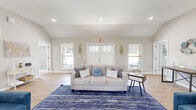 Located at 4727 Grant Line Rd in New Albany, Indiana, this single-floor space offers a clean, modern design with a warm and inviting atmosphere. The layout is open and generously proportioned, allowing natural light to flow freely throughout via multiple windows fitted with blinds.

The main lounge area is a standout feature, showcasing soaring vaulted ceilings that lend a sense of airiness and volume to the room. Light hardwood-style flooring runs throughout the space, complementing the neutral gray and white wall palette. Recessed lighting provides consistent, even illumination across the open floor plan. The lounge is anchored by a large area rug and offers ample room for seating arrangements, with a secondary entertainment area featuring a flat-screen TV on a stylish cabinet, a built-in display niche, and additional seating.

A welcoming entry area with double doors and flanking windows creates a bright, accessible front entrance. The kitchen is well-appointed with white cabinetry, stone countertops, stainless steel appliances, and a window above the sink that brings in natural light. Clean lines and recessed lighting give the kitchen a fresh, functional character.

Overall, the space presents a polished, move-in-ready environment that balances openness with defined functional areas, making it well-suited for a variety of uses.