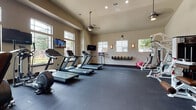 This fitness center is thoughtfully designed to support a comprehensive workout experience. The space features vaulted ceilings that lend an open, airy feel, complemented by a ceiling fan that aids in air circulation during workouts. Neutral-toned walls and dark rubber flooring create a clean, functional aesthetic throughout.

The cardio zone is well-stocked with multiple treadmills and elliptical machines, each positioned near large windows that offer outdoor views and bring in generous natural light. Multiple wall-mounted flat-screen TVs are strategically placed throughout the space, allowing users to stay entertained during their sessions.

The strength training area includes a selection of selectorized weight machines, a free-weight rack with dumbbells, and a dedicated stretching zone equipped with a stability ball and yoga mat storage. A built-in wall-mounted dumbbell rack provides organized storage along one wall.

Wall sconces provide warm accent lighting, while recessed ceiling lights ensure consistent illumination throughout. A large mirror along one wall enhances the sense of space. The fitness center is accessible via a glass-paneled door and benefits from views of the surrounding property through its generously sized windows.