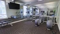 This fitness center is a purpose-built exercise facility designed to support a well-rounded workout routine. The space is outfitted with a selection of cardio and strength training equipment, including elliptical machines and a cable cross-trainer system, providing users with a variety of exercise options. Floor-to-ceiling mirrors line the walls, creating a sense of spaciousness while allowing users to monitor their form during workouts. A wall-mounted flat-screen television adds an entertainment element to exercise sessions. Overhead fluorescent lighting ensures the space is bright and well-illuminated throughout. The flooring consists of a durable, patterned carpet designed to withstand high foot traffic while providing comfort underfoot. A window with blinds allows for natural light to filter into the room, contributing to an open and energizing atmosphere. The layout of the equipment is thoughtfully arranged to allow ample room for movement between machines. Overall, this fitness center offers a clean, functional, and well-maintained environment suited for daily exercise.