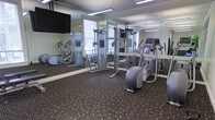 This fitness center is a purpose-built exercise facility designed to support a well-rounded workout routine. The space is outfitted with a selection of cardio and strength training equipment, including elliptical machines and a cable cross-trainer system, providing users with a variety of exercise options. Floor-to-ceiling mirrors line the walls, creating a sense of spaciousness while allowing users to monitor their form during workouts. A wall-mounted flat-screen television adds an entertainment element to exercise sessions. Overhead fluorescent lighting ensures the space is bright and well-illuminated throughout. The flooring consists of a durable, patterned carpet designed to withstand high foot traffic while providing comfort underfoot. A window with blinds allows for natural light to filter into the room, contributing to an open and energizing atmosphere. The layout of the equipment is thoughtfully arranged to allow ample room for movement between machines. Overall, this fitness center offers a clean, functional, and well-maintained environment suited for daily exercise.