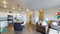 This single-floor unit offers a thoughtfully designed open-concept layout that seamlessly connects the kitchen, dining, and living areas, creating a bright and cohesive living environment. The kitchen is a standout feature, showcasing white cabinetry, stainless steel appliances including a French door refrigerator, and a dark-toned island with granite countertops and bar-height seating — perfect for casual dining or entertaining. Pendant lighting above the island adds a modern touch, complementing the flush-mount ceiling fixtures throughout. Rich, dark hardwood-style flooring runs across the entire space, grounding the interior with warmth and continuity. The living area is generously proportioned, accommodating comfortable seating with ease. Neutral wall tones create a clean, versatile backdrop that adapts well to various décor styles. A dedicated laundry closet with side-by-side washer and dryer adds everyday convenience. Additional hallways suggest the presence of a bedroom and bathroom tucked beyond the main living space. Overall, the layout is efficient yet open, offering a contemporary and functional living experience with well-appointed details at every turn.