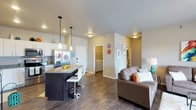 This single-floor unit offers a thoughtfully designed open-concept layout that seamlessly connects the kitchen, dining, and living areas, creating a bright and cohesive living environment. The kitchen is a standout feature, showcasing white cabinetry, stainless steel appliances including a French door refrigerator, and a dark-toned island with granite countertops and bar-height seating — perfect for casual dining or entertaining. Pendant lighting above the island adds a modern touch, complementing the flush-mount ceiling fixtures throughout. Rich, dark hardwood-style flooring runs across the entire space, grounding the interior with warmth and continuity. The living area is generously proportioned, accommodating comfortable seating with ease. Neutral wall tones create a clean, versatile backdrop that adapts well to various décor styles. A dedicated laundry closet with side-by-side washer and dryer adds everyday convenience. Additional hallways suggest the presence of a bedroom and bathroom tucked beyond the main living space. Overall, the layout is efficient yet open, offering a contemporary and functional living experience with well-appointed details at every turn.