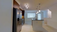 This contemporary 2-floor residence showcases a thoughtfully designed interior with modern finishes throughout. The open-concept kitchen serves as a centerpiece of the main living area, featuring rich warm-toned cabinetry that extends to the ceiling for ample storage. Stainless steel appliances, including a French door refrigerator with water dispenser and a built-in range, lend a polished, professional quality to the space. A central kitchen island with a dark granite countertop and an undermount sink provides additional workspace and casual seating potential, elegantly illuminated by a trio of sleek pendant lights suspended from above. The kitchen flows seamlessly into the living area, which benefits from natural light streaming through multiple windows fitted with white horizontal blinds. Light-toned tile flooring runs throughout the main level, creating a clean and cohesive aesthetic. A track lighting fixture overhead ensures the space remains well-lit. The overall ambiance is modern and refined, combining functional design with contemporary style across both floors, making it a versatile and inviting space.
