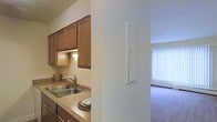 This single-floor residential unit offers a practical and comfortable layout, with the kitchen and living area positioned in close proximity. The kitchen is equipped with warm-toned wooden cabinetry that provides ample storage, complemented by under-cabinet lighting that adds a subtle, functional glow to the workspace. A double-basin stainless steel sink sits beneath the cabinetry, set into laminate countertops that offer a generous prep surface. The kitchen's design is efficient, with a layout that maximizes usability within its footprint.

A structural half-wall separates the kitchen from the adjacent living area, creating a natural division between the two spaces while maintaining an open, connected feel. The living area features wall-to-wall carpeting in a neutral tone, contributing to a cozy atmosphere. A large window dressed with vertical blinds allows natural light to filter into the room, brightening the space and providing a sense of openness. The light-colored walls throughout maintain a clean, neutral aesthetic that offers versatility for personalization.