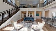 Located at 12 Wilkinson Way in Bluffton, South Carolina, this elegantly appointed clubhouse is designed to impress from the moment you enter. The main level is anchored by a grand, double-height great room with soaring ceilings that draw the eye upward through rows of tall, grid-paned windows, flooding the space with abundant natural light. Warm hardwood-style flooring runs throughout, complementing the neutral, taupe-toned walls and crisp crown molding that frame every room. The open lounge area is thoughtfully furnished with a plush blue velvet sofa, nailhead-trimmed accent chairs, and a rustic wood coffee table arranged atop a layered area rug, creating an inviting setting for gathering. A dramatic iron staircase with a white balustrade ascends to an open mezzanine level, where a conference-style seating area with a large wooden table offers a quiet space for meetings or study, overlooking the main floor below. Adjacent to the lounge, a stylish wet bar serves as a focal point, featuring a striking iridescent mosaic tile accent wall, dark cabinetry, light stone countertops, beverage refrigerators, a coffee station, a wall-mounted television, and bar seating along an extended island. The coffered ceiling detail above the main entrance and an elegant chandelier add refined architectural character throughout.