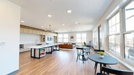 Located at 8681 Edison Plaza Dr in Fishers, Indiana, this single-floor open-plan space offers a thoughtfully designed layout that seamlessly integrates a kitchen, dining, and living area into one cohesive environment. The interior is finished with warm hardwood flooring that runs throughout, complemented by crisp white walls and a prominent wood beam accent that adds architectural character to the space.

The kitchen runs along one wall and features light wood cabinetry, a white quartz countertop, a textured stone tile backsplash, stainless steel appliances including a built-in microwave and compact refrigerator, and an integrated sink. A sculptural multi-globe chandelier anchors the kitchen zone, adding a contemporary design focal point above the large central island table with a white surface and bold dark metal base — ideal for collaborative work or casual dining.

The living area transitions naturally from the kitchen, furnished with a cognac leather sofa, modern lounge chairs, and round ottomans arranged around a statement coffee table. A built-in media wall with open shelving and a wall-mounted TV provides both function and style, framed by large windows that flood the space with natural light and offer views of the surrounding Fishers streetscape. Multiple seating arrangements throughout make this space well-suited for both focused gatherings and informal interaction.