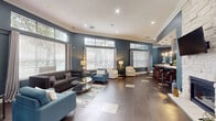 This single-floor space offers a generously sized, open-plan layout that seamlessly integrates a living room, dining area, and kitchen. The living area is defined by deep slate-blue accent walls contrasted against crisp white crown molding and recessed lighting. Large windows fitted with blinds and drape panels flood the space with natural light, while transom windows above add to the impressive ceiling height, enhancing the sense of openness throughout.

The kitchen features a substantial island with a sleek white countertop, dark cabinetry, a stainless steel range hood, built-in appliances, and a textured tile backsplash. Stylish pendant lights hang above the island, which is lined with modern wooden bar stools. The kitchen connects directly to the hallway, which leads through an arched passage to additional rooms.

A dedicated conference room provides a professional setting with two large rectangular tables, leather rolling chairs, wall-mounted televisions set within arched alcoves, and carpeted flooring. Natural light enters through multiple windows dressed with teal curtains. The overall ambiance throughout the space is polished, functional, and contemporary.