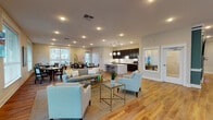 This single-floor community space is designed with an open-concept layout that seamlessly integrates multiple functional zones into one expansive, light-filled environment. Warm hardwood flooring runs throughout, providing a cohesive foundation that ties together each area of the room.

The lounge area serves as a central gathering point, furnished with a plush gray sofa, accent chairs in a light teal patterned fabric, and a glass-top coffee table. Recessed lighting overhead and large windows along the perimeter flood the space with natural light, creating a bright and welcoming atmosphere.

Adjacent to the lounge, a dedicated dining zone features multiple dark wood tables and chairs, well-suited for group gatherings or communal meals. A gallery-style abstract artwork adorns the wall, adding a decorative focal point to the area.

The full kitchen is a standout feature, outfitted with floor-to-ceiling dark espresso cabinetry, light quartz countertops, a white subway tile backsplash, stainless steel appliances, and a large center island with bar seating. Globe pendant lights hang above the island, adding a modern touch.

Beyond the main lounge, the space also includes a fitness area with exercise equipment and a recreation zone featuring a billiards table and wall-mounted televisions, rounding out this well-appointed multi-use community facility.