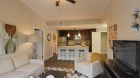 This two-story space showcases a thoughtfully designed open-concept layout that seamlessly connects the living area and kitchen. The kitchen is a standout feature, equipped with rich dark wood cabinetry, granite countertops, and stainless steel appliances including a refrigerator and microwave. A central island with bar seating for three provides both additional workspace and a casual dining option, illuminated by stylish pendant lighting that hangs from the ceiling above. A tile backsplash adds texture and visual interest to the kitchen walls.

The living area flows naturally from the kitchen, offering a comfortable and functional gathering space. Hardwood-style flooring extends throughout, adding warmth and continuity to the open floor plan. A ceiling fan is centrally positioned overhead, enhancing both airflow and the room's aesthetic. Neutral-toned walls complement the overall design, creating a bright and welcoming ambiance. A doorway visible from the main living space hints at additional rooms beyond, while the two-story layout suggests further living space above, making this a well-appointed and versatile home.