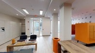 The mail and elevator lobby at 150 Glover Ave in Norwalk, CT is a well-appointed, single-floor common area designed to serve residents with both functionality and contemporary style. The space is bright and open, featuring light wood-look flooring, white structural columns, and a clean, modern aesthetic throughout.

Upon entering through the glass-paneled main doors, residents are welcomed into a spacious lounge area furnished with navy blue chairs and wooden tables, providing a comfortable spot to wait or sort mail. Bold orange accents — including cylindrical waste receptacles, vibrant mailbox units, and accent walls — punctuate the otherwise neutral palette, giving the space a lively, modern character. Typographic wall murals add an artistic touch to the interior.

A dedicated mail area houses a large bank of orange individual mailboxes alongside an extensive wall of smart package lockers, offering secure and convenient parcel retrieval. Skylights and recessed lighting ensure the space remains well-lit throughout the day.

The elevator alcove is distinctly finished with deep teal-blue walls and houses two stainless steel elevator doors, creating a striking visual contrast to the brighter main lobby. Orange ottomans nearby provide additional casual seating. Overall, the lobby delivers a cohesive, energetic design that balances practical resident services with an inviting communal atmosphere.