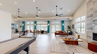 This generously sized single-floor community living space offers an open, airy layout designed for both relaxation and recreation. The room is anchored by soaring ceilings with dual large-blade ceiling fans that enhance airflow throughout the expansive floor plan. Light hardwood flooring runs continuously underfoot, complementing the bright, neutral color palette accented by bold teal structural columns that add a modern, vibrant touch.

A full-size pool table takes center stage in the main area, accompanied by a wall-mounted cue rack and a large flat-screen television — making it an ideal entertainment hub. A dedicated shelf unit displays board games and recreational accessories, adding a communal, welcoming character. Adjacent to the game area, a cozy lounge zone features a rich brown leather sofa, plush area rug, and a striking floor-to-ceiling tiled fireplace surround that serves as a dramatic architectural focal point.

On the opposite end, a dining and bar-height seating area provides space for gathering, bathed in natural light streaming through multiple large windows and glass-paned French doors that frame outdoor views. Recessed lighting and wall sconces throughout ensure warm, even illumination. The overall ambiance is modern and inviting, blending comfort with a lively, social atmosphere.