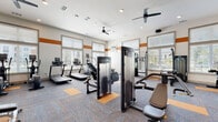 This spacious fitness center is thoughtfully designed to support a full range of workout styles. The interior is defined by soaring ceilings accented with a bold orange horizontal stripe that runs along the upper walls, creating a dynamic and energetic atmosphere. The flooring consists of patterned carpet tiles in grey and orange, adding visual interest while providing cushioning underfoot.

Natural light floods the space through numerous large windows fitted with horizontal blinds, offering views of the surrounding exterior. Ceiling fans are installed throughout to maintain airflow and comfort during workouts. Recessed lighting and wall-mounted speakers further enhance the environment.

The cardio zone houses multiple treadmills, elliptical trainers, a stair climber, and rowing machines. The strength training area features a cable machine, multi-functional weight stations, and additional resistance equipment. A free weights section includes a full rack of dumbbells and adjustable workout benches positioned in front of a large wall mirror, with wall-mounted TVs nearby for entertainment.

Near the entrance, a dedicated functional training station offers stability balls, medicine balls, kettlebells, resistance bands, and foam rollers neatly organized on storage racks. Double glass doors provide easy access, completing this well-appointed and comprehensive fitness facility.