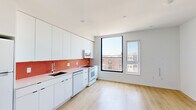 This thoughtfully designed one-bedroom, one-bathroom residence offers an efficient and modern living experience across a single floor. Light-toned hardwood floors run throughout, lending warmth and visual continuity to every room.

The kitchen is a standout feature, showcasing sleek flat-front white cabinetry extending from floor to ceiling for ample storage. A vibrant red penny-tile backsplash adds a bold pop of color and personality, complemented by matching countertops and a full suite of white appliances including a refrigerator, dishwasher, range, and over-the-range microwave. Large black-framed floor-to-ceiling windows flood the kitchen and living area with natural light and frame open urban rooftop views.

The bedroom is a calm, minimalist retreat with its own generous black-framed windows offering additional city views and natural light. A wall-mounted mini-split air conditioning unit ensures comfort year-round, and a built-in closet provides practical storage.

The bathroom continues the design's signature use of color, featuring a soaking tub-shower combination surrounded by a striking mosaic tile pattern of red and gray squares, creating an eye-catching, artistic focal point. The overall aesthetic of the unit is clean, contemporary, and cohesive, with a consistent palette of white walls and warm wood flooring throughout.