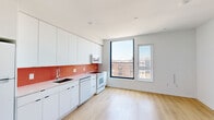 This thoughtfully designed one-bedroom, one-bathroom residence offers an efficient and modern living experience across a single floor. Light-toned hardwood floors run throughout, lending warmth and visual continuity to every room.

The kitchen is a standout feature, showcasing sleek flat-front white cabinetry extending from floor to ceiling for ample storage. A vibrant red penny-tile backsplash adds a bold pop of color and personality, complemented by matching countertops and a full suite of white appliances including a refrigerator, dishwasher, range, and over-the-range microwave. Large black-framed floor-to-ceiling windows flood the kitchen and living area with natural light and frame open urban rooftop views.

The bedroom is a calm, minimalist retreat with its own generous black-framed windows offering additional city views and natural light. A wall-mounted mini-split air conditioning unit ensures comfort year-round, and a built-in closet provides practical storage.

The bathroom continues the design's signature use of color, featuring a soaking tub-shower combination surrounded by a striking mosaic tile pattern of red and gray squares, creating an eye-catching, artistic focal point. The overall aesthetic of the unit is clean, contemporary, and cohesive, with a consistent palette of white walls and warm wood flooring throughout.