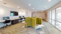 This contemporary single-floor workspace is thoughtfully designed to support a range of collaborative and focused work activities. The main open area is anchored by a cluster of distinctive chartreuse-upholstered pod seating units arranged back-to-back, offering semi-private nooks ideal for informal meetings or individual focus work, each equipped with compact side tables. Along one wall, a built-in workstation counter hosts multiple desktop computers beneath a large wall-mounted display, providing a dedicated technology hub. Recessed lighting throughout keeps the space bright and welcoming, complemented by an eye-catching patterned carpet in muted gray and yellow tones that ties the design together.

A glass-walled private meeting room off the main area features a round table with sleek black leather chairs and a large arched window overlooking an outdoor courtyard, filling the room with natural light and pleasant views. A second, more enclosed conference room accommodates a rectangular white table with upholstered rolling chairs, offering a quieter setting for focused discussions. Curated abstract artwork is displayed throughout, adding visual interest to the clean white walls. Built-in shelving and additional workstation areas round out the space, making it a well-equipped, versatile environment suited for collaboration, meetings, and productivity.
