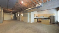 This single-floor space showcases a distinctive industrial loft aesthetic defined by its exposed concrete and brick ceiling structure, prominent ductwork running overhead, and an open, expansive floor plan. The generous main living area is finished with neutral wall-to-wall carpeting, creating a warm contrast against the raw, utilitarian ceiling elements above. Structural columns punctuate the space, adding to the urban character while subtly defining different zones within the open layout.

The kitchen is positioned along one wall and features light maple cabinetry with dark granite-style countertops, providing a clean and functional workspace. Stainless steel appliances, including a gas range and microwave, are integrated into the cabinetry, offering a modern touch. The kitchen flooring transitions to a hard surface, distinguishing it from the carpeted living area.

Track lighting and recessed fixtures are mounted to the exposed ceiling, ensuring the space is well-lit throughout. Additional rooms or utility areas are visible toward the rear of the unit, accessible via open doorways. The overall ambiance blends raw industrial elements with clean, contemporary finishes, making this a versatile and visually compelling space suited to a variety of uses.