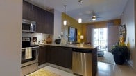 This single-floor space showcases an open-concept layout that seamlessly connects the kitchen and living area, creating a bright and functional environment. The kitchen is outfitted with dark espresso cabinetry that contrasts elegantly with granite countertops, providing ample prep and storage space. Stainless steel appliances, including a dishwasher, microwave, and range, lend a contemporary, polished look. A peninsula with an undermount sink extends the counter space and doubles as a casual dining or entertaining area, illuminated by modern pendant lighting that hangs at varying heights for visual interest. The backsplash adds a clean, refined touch beneath the upper cabinetry. The adjoining living area benefits from a ceiling fan, accent lighting, and a large flat-screen television mounted on a warm yellow-toned accent wall, adding a bold pop of color to the space. Natural light flows generously through sliding glass doors that open to a balcony, offering views of the surrounding cityscape. Neutral tile flooring runs throughout, accented by a patterned area rug that adds warmth. The overall ambiance is modern and inviting, with a cohesive blend of dark tones and warm accents.
