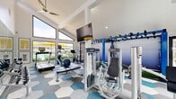 Located at 8114 W Britton Rd in Oklahoma City, Oklahoma, this fitness center offers a thoughtfully designed exercise space filled with an impressive array of equipment and modern amenities. The room is defined by its striking vaulted, cathedral-style ceilings that soar overhead, creating an open and energizing atmosphere. Large, floor-to-ceiling angled windows along one wall flood the space with abundant natural light while offering views of an outdoor pool area, complete with yellow umbrellas, adding a vibrant backdrop to workouts. A ceiling fan and wall-mounted flat-screen television enhance comfort and entertainment during exercise sessions.

The flooring features a bold geometric hexagonal tile pattern in shades of blue, gray, and white, lending the space a contemporary, polished aesthetic. Equipment includes Life Fitness cardio machines such as treadmills, ellipticals, and a stationary bike, along with multiple strength training machines and a free-weight bench. A full dumbbell rack is positioned in front of a large wall mirror, maximizing functional use of the space.

A dedicated functional training zone features a bold blue multi-purpose rig with monkey bars, suspension straps, and resistance band attachments, set over an artificial turf mat with battle ropes — ideal for dynamic, high-intensity workouts. Large sliding glass doors provide easy access and additional natural light, completing this well-rounded and inviting fitness facility.