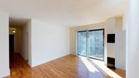 This single-floor space offers a clean, minimalist aesthetic defined by warm hardwood flooring, crisp white walls, and a textured ceiling that adds subtle character overhead. The living area is generously proportioned, bathed in abundant natural light that streams in through a large sliding glass door fitted with vertical blinds. The door opens onto a private balcony, providing an outdoor extension of the living space and framing views of the surrounding urban streetscape. The open layout creates a versatile living environment well-suited to a variety of furniture arrangements. The floor-to-ceiling sliding glass door assembly serves as both a functional entryway to the balcony and a striking architectural focal point, drawing the eye outward and reinforcing the connection between interior comfort and the city beyond. The warm tones of the hardwood floors contrast neatly with the neutral white walls, lending the space a bright and airy quality throughout. Overall, the unit presents a straightforward yet appealing design that prioritizes natural light, open space, and easy access to the outdoors.