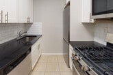 Located at 16-70 Bell Blvd in the Bayside neighborhood of Queens, NY, this single-floor residence offers a clean, well-maintained living environment with consistent design details throughout. The living area is anchored by warm parquet wood flooring — a hallmark of the space — and is framed by crisp crown molding and soft neutral-gray walls. Large sliding glass doors open to a private balcony, flooding the room with natural light and providing views of the surrounding neighborhood. The bedroom continues the parquet flooring theme and features two generous built-in closets, offering ample storage. A second room, also with parquet flooring and a window overlooking the street, provides versatile additional living space. The galley-style kitchen is efficiently designed with white shaker cabinetry, dark stone countertops, a white subway tile backsplash, light tile flooring, and stainless steel appliances including a gas range and refrigerator. The bathroom is tastefully finished with large-format beige wall tiles, a decorative mosaic tile accent border, a white vanity with mirrored medicine cabinet, and grab bar accessibility features. Together, these elements create a tidy, functional, and comfortable urban residence in a well-established Queens community.