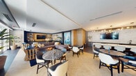 Located at 1000 Seminole Drive in Fort Lauderdale, FL, this elegantly appointed single-floor lounge space offers a sophisticated blend of contemporary design and coastal-inspired aesthetics. The open-plan layout accommodates a variety of seating arrangements, seamlessly flowing from one zone to the next across herringbone-patterned light wood flooring.

The main gathering area features a generous sectional sofa paired with a dark marble-topped coffee table and an eclectic mix of upholstered chairs in cream, navy, and gold tones. A large nautical painting serves as a dramatic focal point, complementing the coastal theme carried throughout the space. Adjacent dining clusters with round marble tables and stylish chairs provide intimate settings for smaller groups.

A dedicated media lounge area includes built-in cabinetry finished in dark wood, floating shelves displaying curated coastal décor, and a wall-mounted flat-screen television. On the opposite end, a bar or service counter sits beneath a striking marble accent wall, illuminated by a modern multi-arm chandelier.

Floor-to-ceiling glass doors and expansive windows flood the interior with natural light while providing direct visual and physical access to a landscaped outdoor terrace with palm trees and lounge seating. Lush potted tropical plants are thoughtfully placed throughout, reinforcing the warm, resort-like ambiance of this inviting communal space.