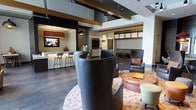 This single-floor communal space showcases a thoughtfully designed interior that blends modern aesthetics with functional versatility. Exposed wooden ceiling beams add architectural character against a clean white ceiling, while dark hardwood-style flooring grounds the space with warmth and contrast.

The layout is organized into several distinct zones, each serving a different social purpose. A bar-height counter with sleek barstools offers a casual perch near a wall-mounted television, making it ideal for informal gatherings. Adjacent to this, a lounge area features a dramatic curved high-back chair as a focal point, surrounded by an eclectic mix of seating including leather accent chairs and upholstered pieces arranged on a patterned area rug.

Further into the space, a booth-style banquette with textured upholstery provides a more intimate setting, while recessed and pendant lighting throughout creates layers of illumination that enhance the overall ambiance. A secondary wall-mounted screen is integrated into a neutral display niche, adding a media-ready element to the environment.

Overall, the space presents a polished, contemporary atmosphere well-suited for socializing, relaxing, or casual work.