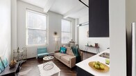 This single-floor loft-style unit showcases an industrial-modern aesthetic defined by dramatically high ceilings with exposed structural beams and visible ductwork, creating an airy, open ambiance throughout. Rich, dark hardwood flooring runs continuously across the main living areas, anchoring the space with warmth and contrast.

The open-concept living area is generously proportioned, featuring tall windows that flood the space with natural light. The kitchen is well-appointed with dark espresso cabinetry extending to ceiling height, white quartz countertops, stainless steel appliances including a range with hood vent, dishwasher, and refrigerator, along with a stacked in-unit washer and dryer located down a connecting hallway.

The bedroom offers an intimate atmosphere with soaring ceilings continuing from the main space. A unique dual-closet configuration with dark-framed doors provides ample organized storage. The bathroom is clean and functional, featuring a dark vanity with white countertop, a large mirror, and a tub-shower combination. Throughout the unit, the interplay of white walls, dark finishes, and industrial ceiling details creates a cohesive and contemporary loft character.