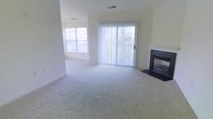 This single-floor space offers a clean, open layout with a welcoming and airy atmosphere throughout. The main living area is generously sized, finished with plush wall-to-wall carpeting that adds warmth and comfort underfoot. Neutral tones across the walls and flooring create a bright, versatile backdrop suited to a variety of interior styles.

A standout feature of the living area is the sleek fireplace framed in dark tile, providing a refined focal point and a cozy gathering spot. Natural light pours into the space through multiple windows, including a large sliding glass door dressed with vertical blinds that leads to an outdoor area, offering views of the surrounding environment and easy access to the exterior.

The open flow of the layout enhances the sense of spaciousness, with the living zone seamlessly connecting to adjacent areas. Recessed ceiling vents indicate a central air and heating system for year-round climate comfort. The overall ambiance is light, neutral, and move-in ready, with the combination of soft carpeting, abundant natural light, and the warmth of the fireplace making this a practical and inviting living space.