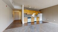 This single-floor unit is designed around an open-concept layout that seamlessly connects the kitchen and living areas. The kitchen occupies a defined zone along one wall, featuring warm honey-toned wood cabinetry that extends to the upper and lower sections, offering generous storage. Light-toned countertops run the length of the kitchen, complemented by a full suite of white appliances, including a coil-burner range with an overhead exhaust hood, a dishwasher, and a refrigerator. A track lighting system overhead provides focused illumination throughout the cooking space. The flooring transitions from wood-look hard flooring in the kitchen to soft, neutral-toned carpet in the open living area, which is a notably spacious room bathed in natural light from large, multi-pane windows. The neutral wall tones throughout contribute to a clean, understated aesthetic. A textured ceiling adds a subtle design element across the main living spaces. The bathroom is well-appointed with a full soaking tub and shower combination, a wood-toned vanity with a white countertop, wood-look flooring, and chrome fixtures. The entry area provides a straightforward transition into the main living space, with a closet nearby for additional storage.