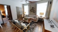 This single-floor living space showcases a thoughtfully designed interior that balances comfort and style. The living area serves as the focal point, featuring rich hardwood flooring that anchors the room with warmth and character. Neutral gray-toned walls provide a versatile backdrop, complemented by earthy accents throughout.

The seating arrangement is both functional and inviting, centered around a tufted linen sofa adorned with decorative throw pillows and a cozy blanket. Two wooden-framed armchairs with gray upholstery face the sofa, creating an intimate conversation area. A wooden coffee tray table sits atop a plush shag area rug, adding texture and visual interest to the space.

Natural light filters in through multiple windows dressed with grayish curtains and blinds, brightening the room without compromising privacy. A ceiling fan overhead ensures year-round comfort. Decorative circular metallic wall accents add an artistic touch above the sofa, while table lamps on either side of the room provide warm ambient lighting. A six-panel door near the entryway completes the space, contributing to its clean, cohesive aesthetic.