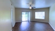 This single-story residence offers a well-designed layout that balances comfort and functionality. The main living area immediately draws attention with its rich, dark hardwood flooring that runs throughout, reflecting natural light and adding warmth to the space. A ceiling fan with a built-in light fixture is centrally positioned overhead, providing both airflow and ambient illumination beneath a textured ceiling. The room is generously lit by two large windows fitted with horizontal blinds, as well as a sliding glass door dressed with sheer vertical drapes. The sliding door opens directly to an outdoor area, allowing views of lush greenery and seamlessly connecting the interior to the exterior. Neutral-toned walls provide a clean, versatile backdrop that complements a wide range of furnishing styles. White baseboards offer a crisp contrast to the flooring and wall tones, contributing to a tidy, finished appearance throughout. The open feel of the living space, combined with the multiple light sources and outdoor access, creates a bright and airy atmosphere well-suited for everyday living and entertaining alike.