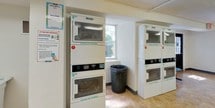 This shared laundry facility is designed with practicality and efficiency in mind, providing residents with a well-equipped space for their laundry needs. The room features a row of commercial-grade front-loading washing machines lining one wall, complemented by several stacked dryer units by Hercules positioned throughout the space. The machines are numbered for easy identification, and usage instructions are clearly posted on the walls for resident reference.

The facility is finished with durable tile flooring throughout, providing an easy-to-clean surface that withstands heavy foot traffic. White walls and fluorescent overhead lighting keep the space bright and functional. Natural light filters in through windows along one wall, offering views of surrounding greenery and adding a welcoming element to the utilitarian environment.

A small seating area with two blue chairs is available for residents waiting on their laundry. The entry area includes a payment kiosk, a fire extinguisher, and an exit sign, reflecting the facility's attention to safety and convenience. A folding table provides a practical surface for handling laundry. The overall layout is open and uncluttered, ensuring residents can navigate the space with ease.
